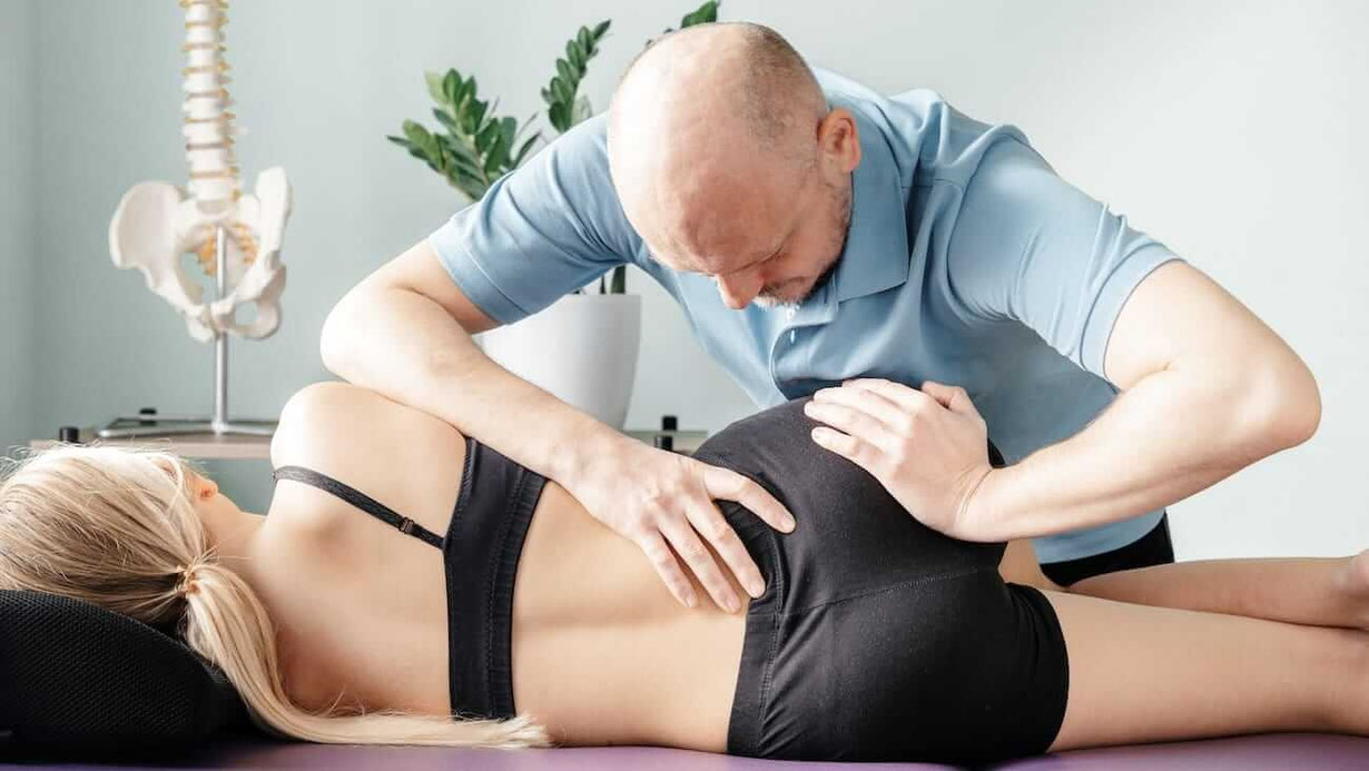Faszien Therapeut beugt sich über eine Behandlungsliege und arbeitet mit den Händen am Becken einer liegenden Frau.