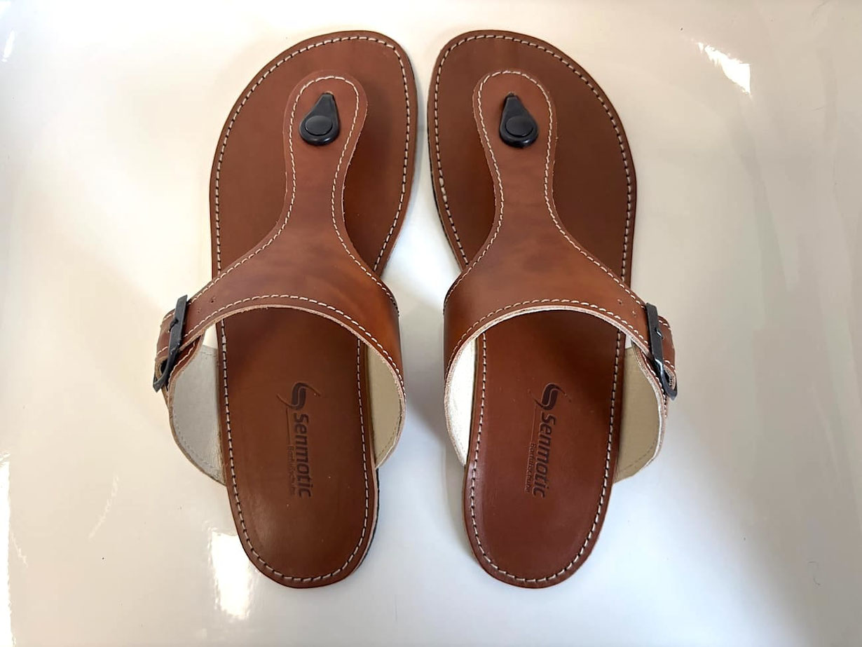 Handgefertigte Flip-Flops aus braunem pflanzlich gegerbten Leder mit Nähten in Weiß. Mit eingestanzten Senmotic Logo. Verstellbar mit Schnallen aus Metall. Laufsohle 3 mm Kautschuk in Schwarz.