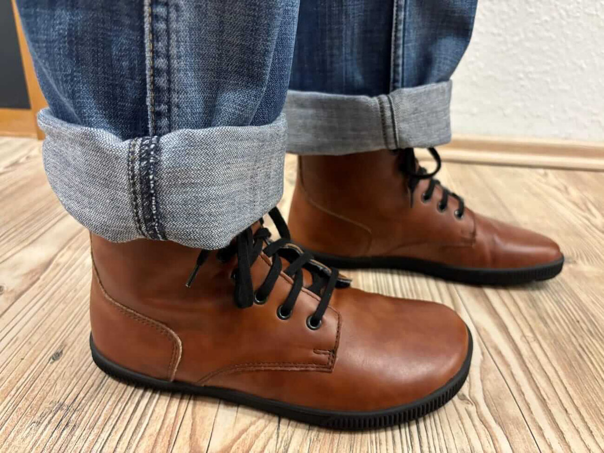 Winterschuh Barfuß Modell Blizzard in braunem Leder mit schwarzen Lammfell an den Füßen einer Person die Jeans trägt.