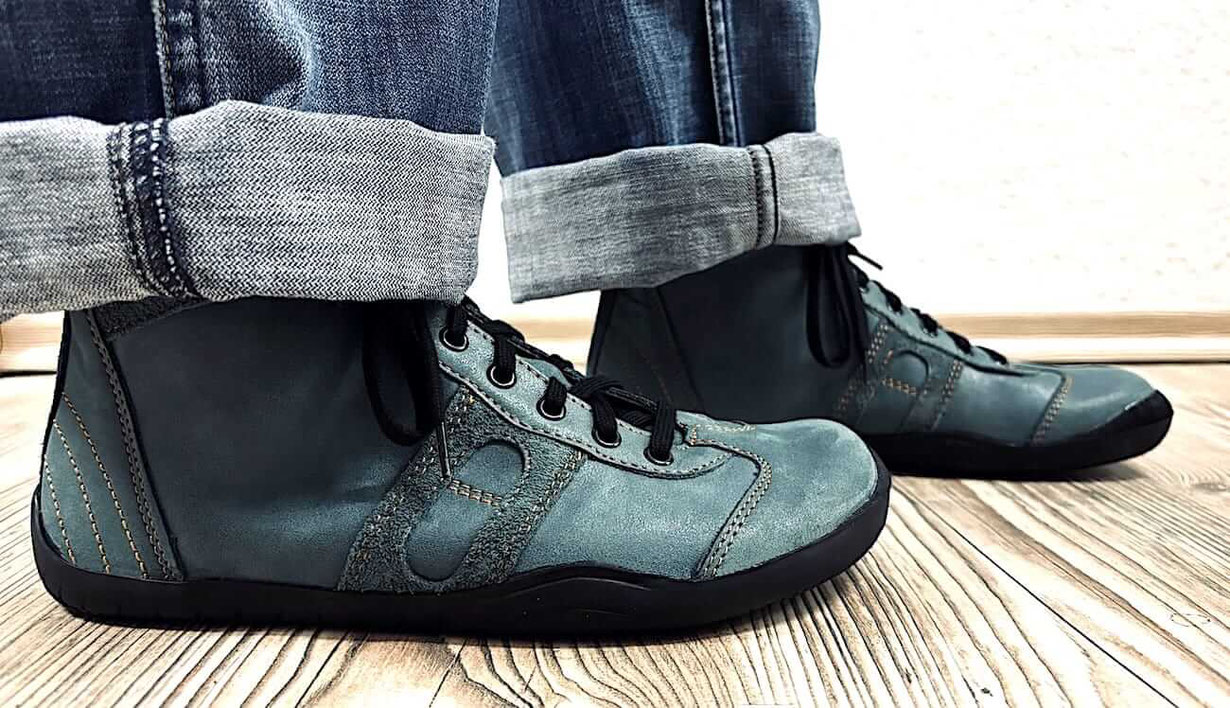Leder Barfußschuhe Modell Retro in der Lederfarbe Petrol mit orangen Nähten an den Füßen einer Frau, die Jeans dazu trägt.