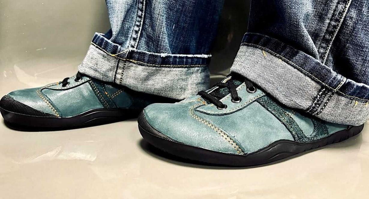 Senmotic Barfußschuhe Modell Diavolo Lederfarbe Petrol Nähte Orange an den Füßen einer Person mit Jeans