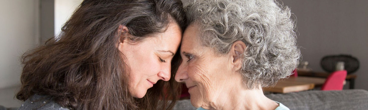 Zwei Frauen unterschiedlicher Generationen sitzen Stirn an Stirn auf dem Sofa, Nähe und tiefe Verbundenheit zwischen Alt und Jung sichtbar.
