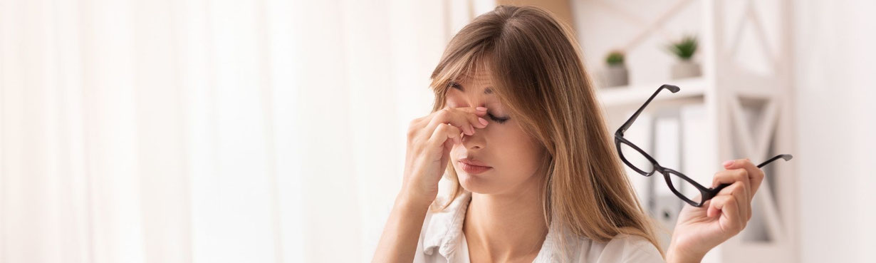 Eine erschöpfte Frau sitzt am Schreibtisch, nimmt die Brille ab und hält sich die Augenpartie, während vor ihr ein Laptop steht – ein stiller Moment von Überforderung und innerer Müdigkeit.