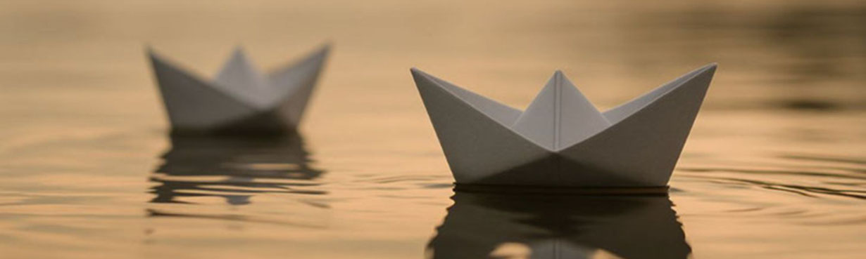 Zwei weiße Papierboote treiben auf ruhigem Wasser in warmem Abendlicht. Das vordere Boot ist scharf fokussiert, das hintere verschwommen im Hintergrund. Die sanften Wellen spiegeln das goldene Licht, die Szene wirkt still, poetisch und kontemplativ.