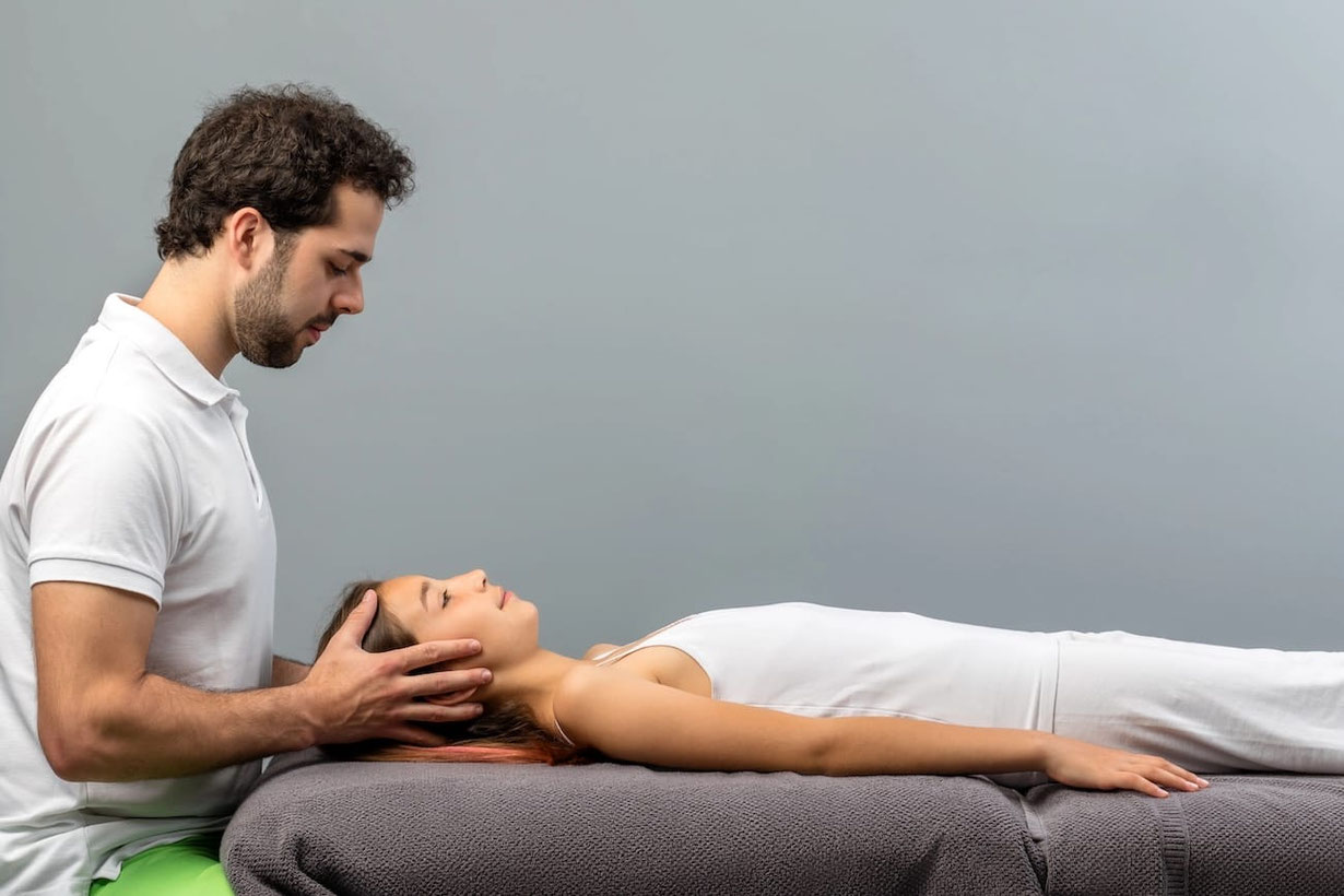 Sitzender Senmotic Faszientherapeut behandelt den Kopf einer liegenden Patientin 