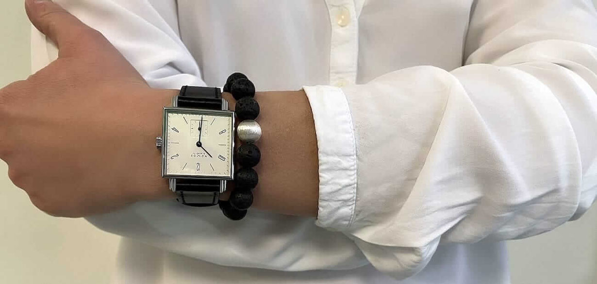 Linkes Handgelenk einer Frau mit einer quadratischen Uhr und einem schwarzen Lava Perlenarmband mit silberner Perle