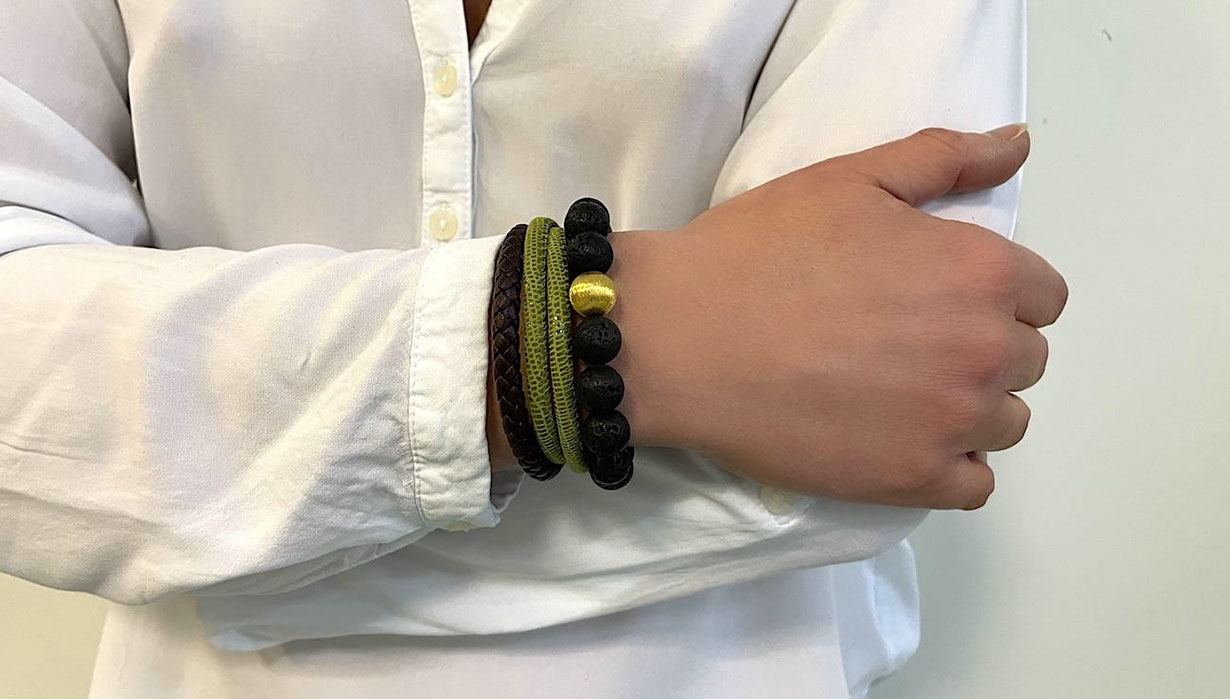Eine Frau mit verschränkten Armen trägt am Handgelenk ein einfaches schwarzes Lederarmband. Ein weiteres doppelt gewickeltes grünes Lederarmband und ein Perlenarmband aus schwarzen Lavaperlen mit einer vergoldeten Perle