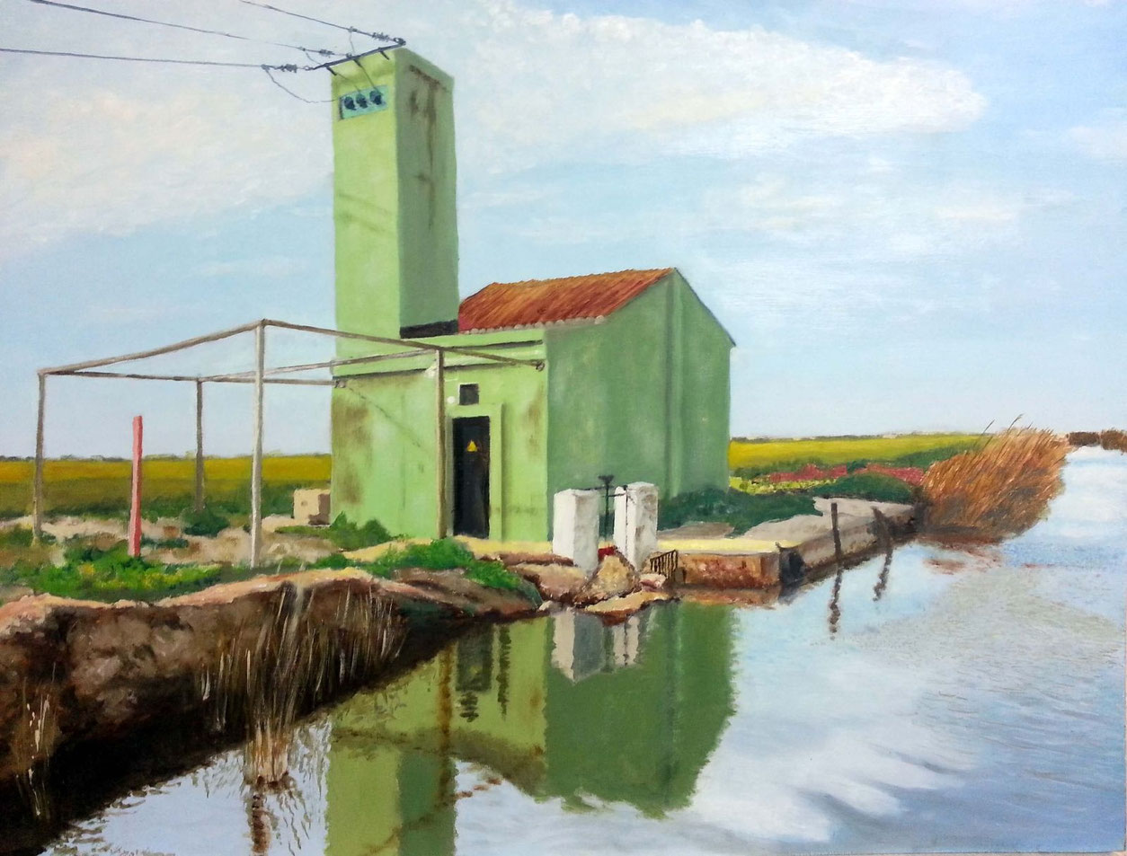Edificio verde junto a un canal, con torre estrecha, reflejo en el agua y paisaje agrícola alrededor.