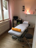 Behandlungsraum mit verstellbarer Liege, grauer Abdeckung, orangefarbenem Nackenrollen-Kissen und schwarzem Hocker vor Fenster mit rosa Vorhang