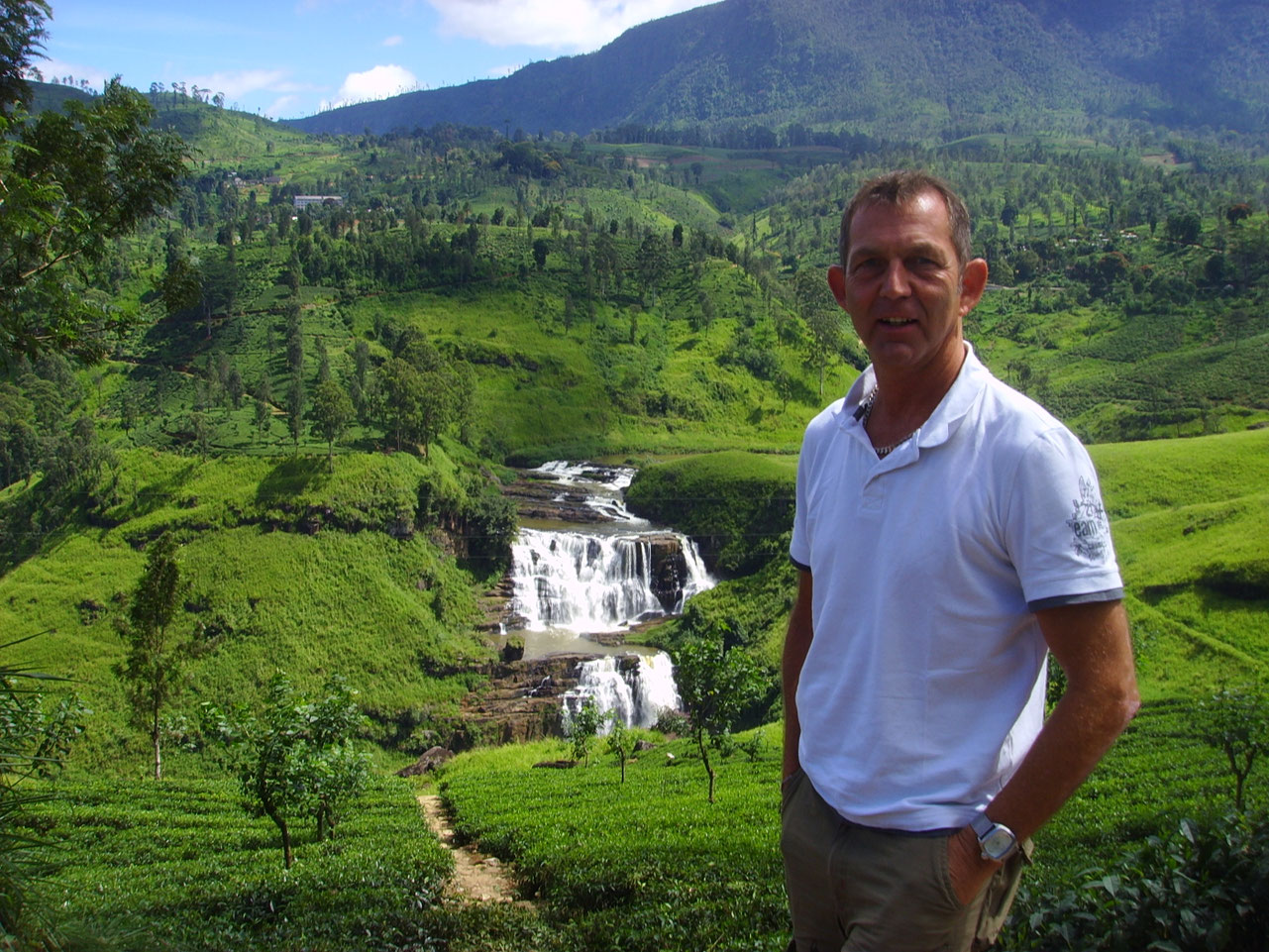 Sri Lanka Reiseexperte Olaf Diroll steht vor einem Wasserfall und einer Teeplantage im Hochland von Sri Lanka und prüft die Qualität der angebotenen Rundreisen