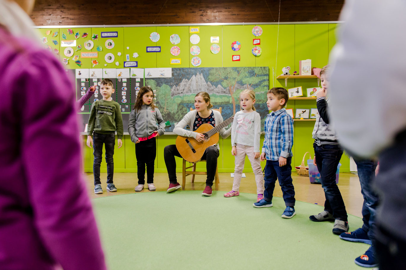 Kinder_stehen_im_Kreis_mit_Lehrperson_an_Gitarre