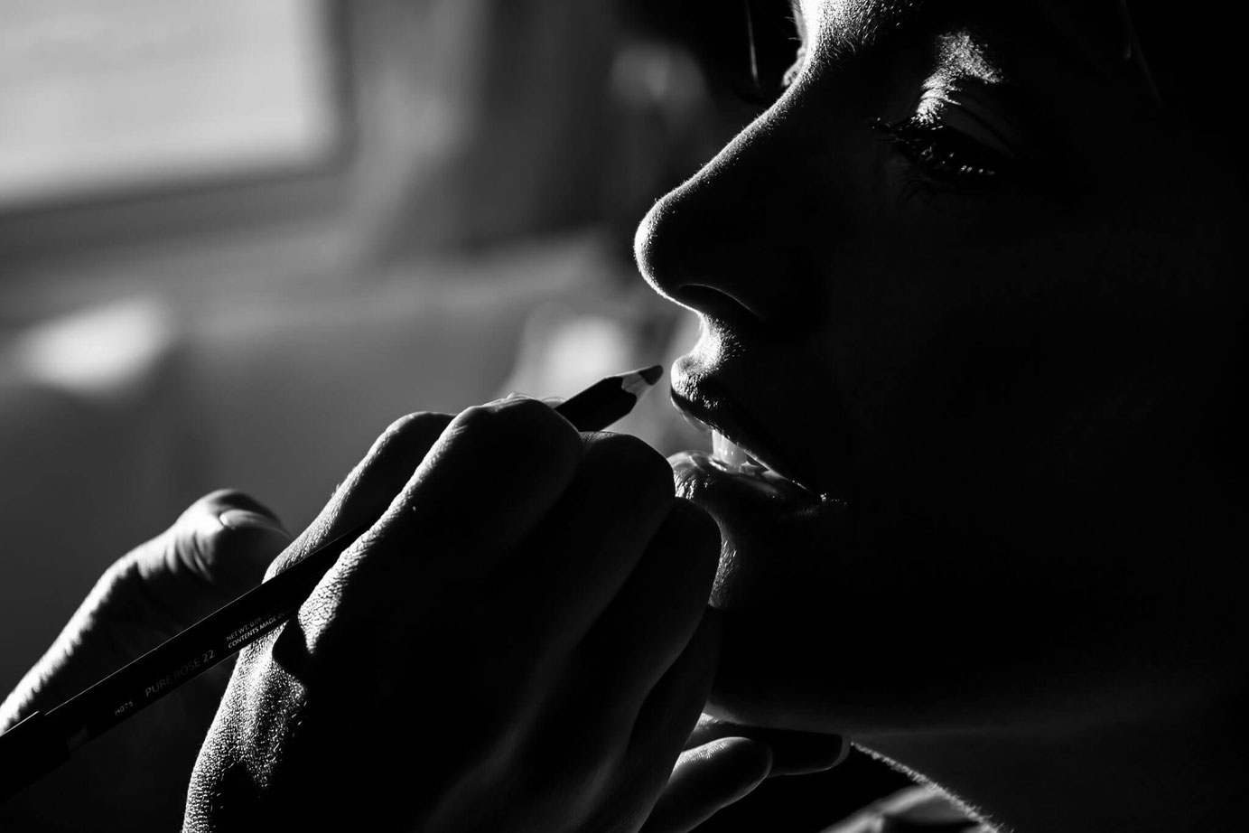 Black and white photo of make-up artist applying lip liner to bride. Photo is cropped to show only make-up artist's hand and bride's face. Light behind bride is visible around rim of bride's face. 