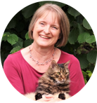 Lachende Astrid Huber mit magentafarbenen T-Shirt und braun-schwarzer Tigerkater Rico auf ihrem Schoß. Grünpflanzen im Hintergrund.