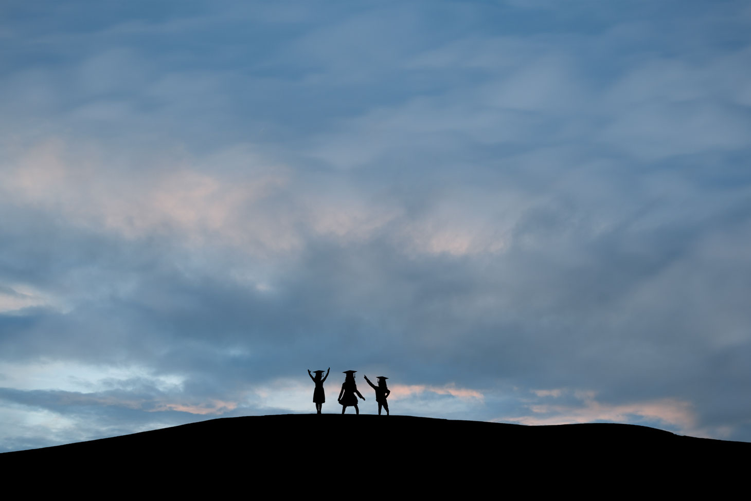 silhouettes of 3 girls in grad caps on a hill against a blue sky with bits of pink