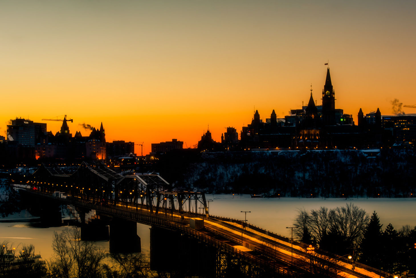 Photo of Ottawa parliament taken at sunrise from Gatineau Quebec. Chateau Laurier and bridge between Ontario and Quebec are also visible. River under bridge is frozen. 