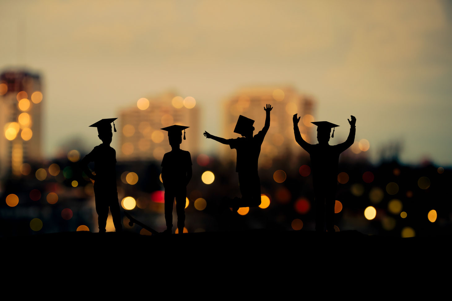 silhouettes of 4 boys in grad caps in front of a city view at sunset.