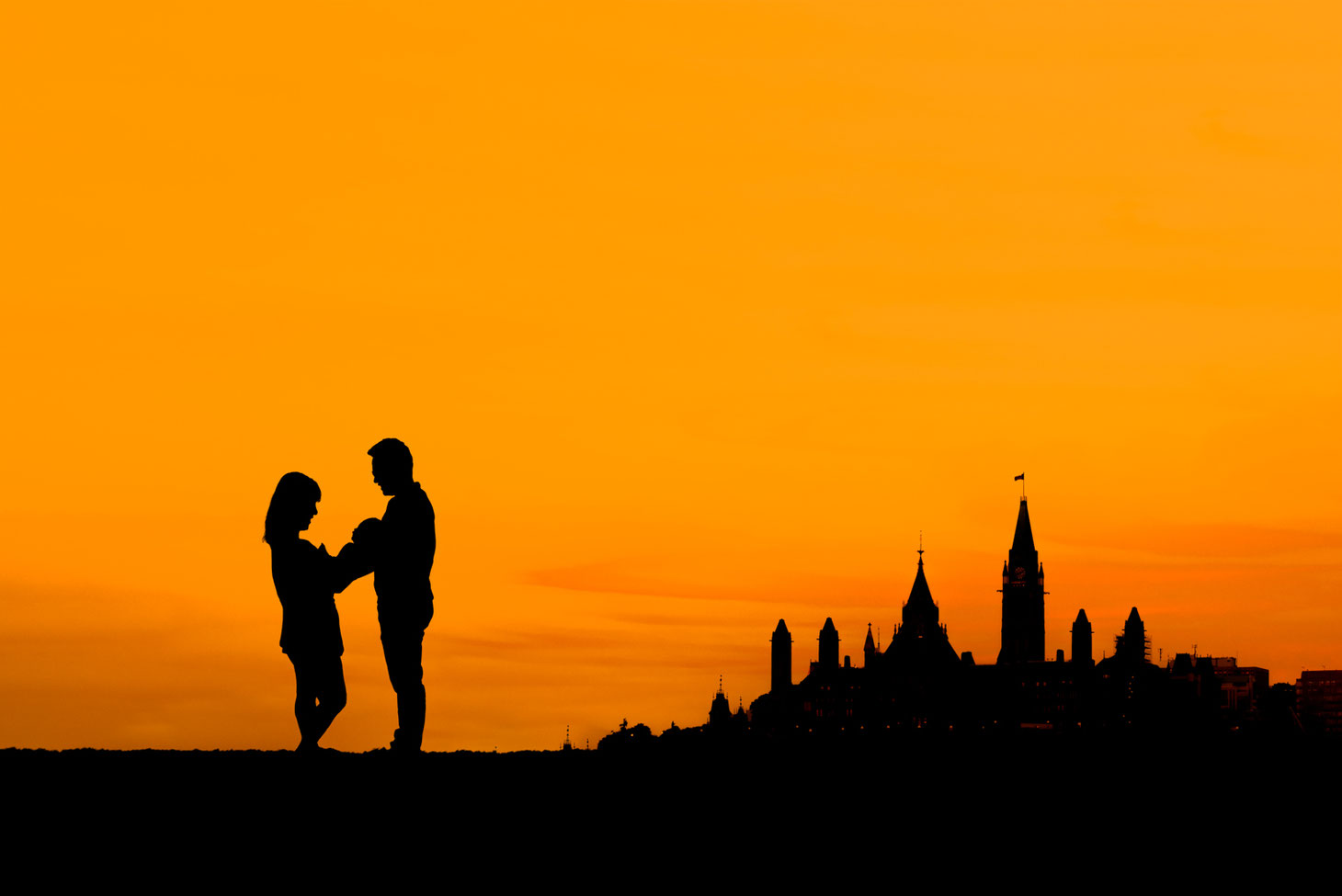  silhouette of mother, new baby, and father next to a silhouette of parliament hill in Ottawa Ontario. Sky is orange with a small amount of clouds. 