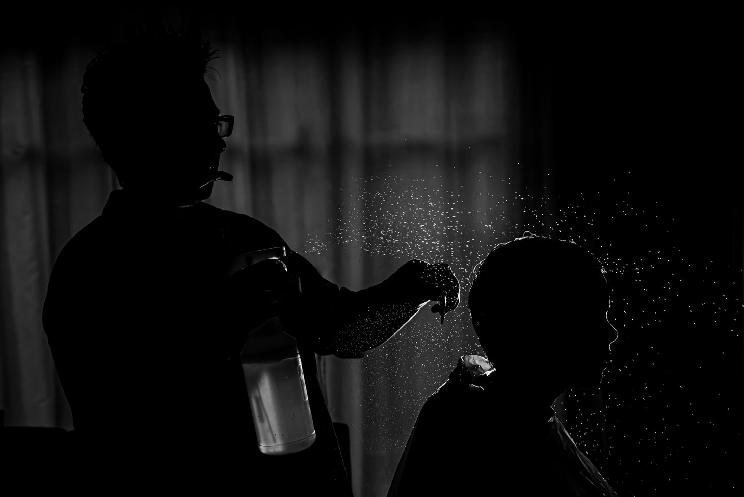 Black and white photo of mom giving child a haircut. Photo is backlit with child and mom in shadow and water droplets visible