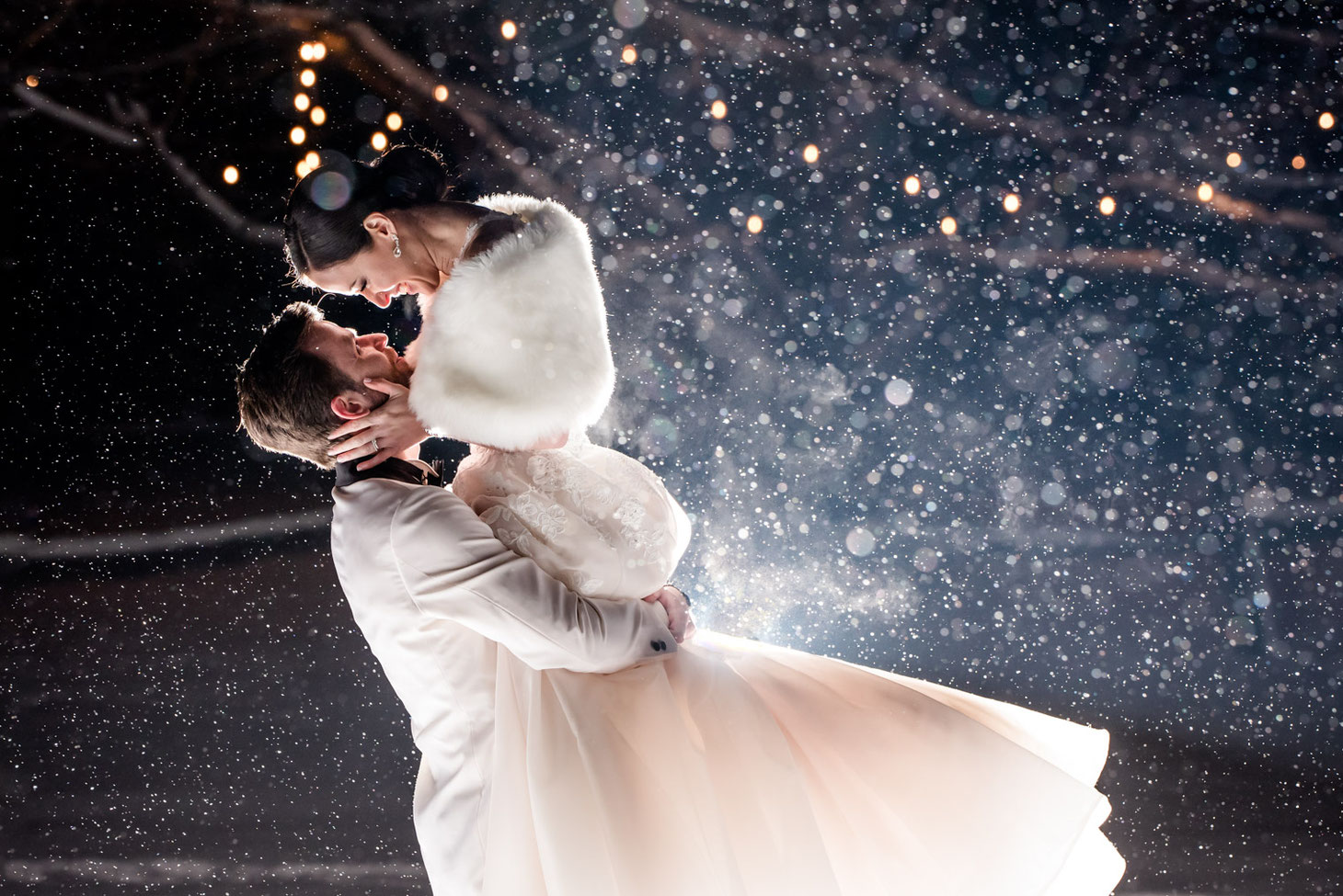 Bride and groom backlit on a snowy evening. Groom is in a white coat and is lifting bride in the air. Bride is in a light pink dress with a white shall.