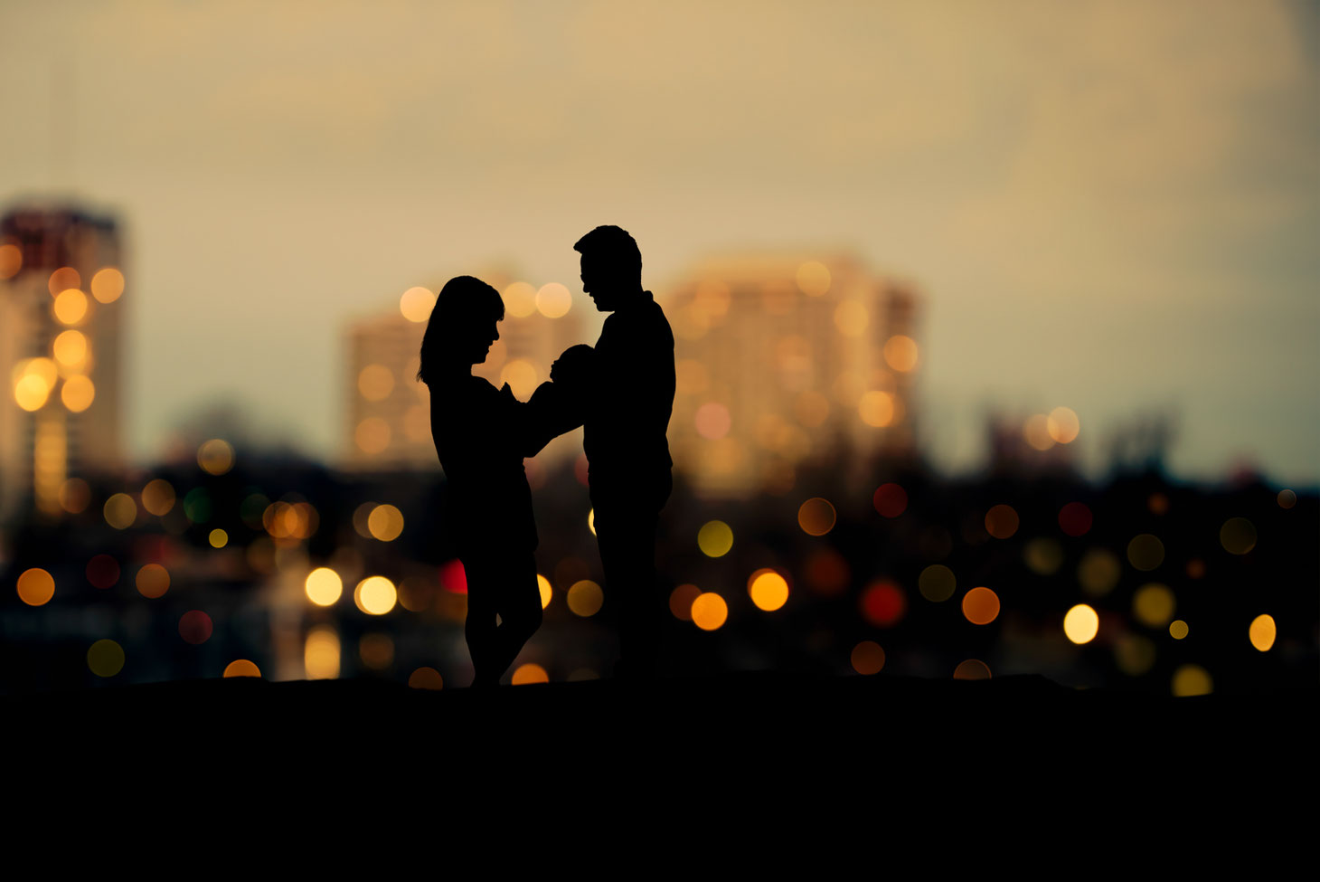 silhouette of mother, new baby, and father against bokeh city view at sunrise.
