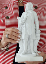 Eine Hand mit einem Ring und glitzernden silbernen Fingernägeln hält eine Bach-Statue vor das rot/weiß gestreiftes Jackett der Trägerin. Die andere Hand unterstützt.