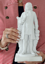 One hand with a ring and glittering silver fingernails holds a statue of Bach in front of the wearer's red and white striped jacket. The other hand supports.