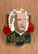 A Bach badge is lying on a wooden base, behind Bach is a red decorative field, "Bach" is written in large letters on his jacket. The badge is about the size of two thumbs.