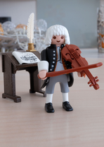The Little Bach Figure. It is standing on a piece of furniture and has a violin in its hand. Behind Bach is a standing desk with a pen, inkwell and music book.