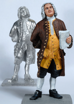 Two fully sculpted, identical tin figures of Bach stand diagonally offset one behind the other. Bach is looking upwards and holding a sheet of music in his hand. The figure in front is painted, the one behind is unpainted. Both stand on a pedestal.