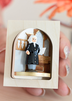 A barely visible hand holds a small box with a cutout for a turntable. On it, you can see Bach in front of the pipe organ. The box is very small.