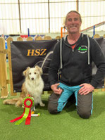 Wolfgang kniet neben seinem Hund Sammy, der vor einem Banner sitzt und eine rote und grüne Schleife vor sich hat.