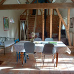 Séjour avec table à manger, poutres apparentes, mur en briques, escalier vers la mezzanine et poêle à bois en arrière-plan