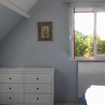 Chambre mansardée avec commode blanche, petite table ronde et fenêtre donnant sur les arbres