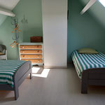 Chambre mansardée avec deux lits simples rayés vert et blanc, commode et fauteuil en osier