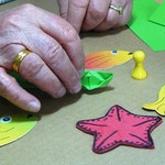 Ältere Patientin hält ein kleines gefaltetes Papier-Boot. Daneben bunte Papierfiguren.