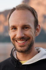 Ein Portrait von Timon Kunter, Osteopath in der Praxis Octopus in Graz. Das Foto zeigt ihn vor einem See, mit dem Traunsee unscharf im Hintergrund. Er lächelt und hat einen schwarzen Pullover an.