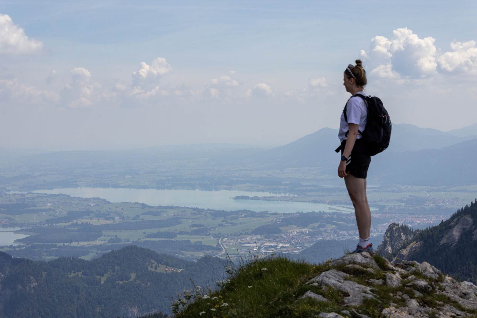 Eine Wanderin genießt den Panoramablick über Seen, Berge und das Alpenvorland von einem Gipfel oberhalb des Tannheimer Tals. Nach der Wanderung zur Bad Kissinger Hütte lädt das Wellnesshotel DER ENGEL zum Entspannen im Spa ein.