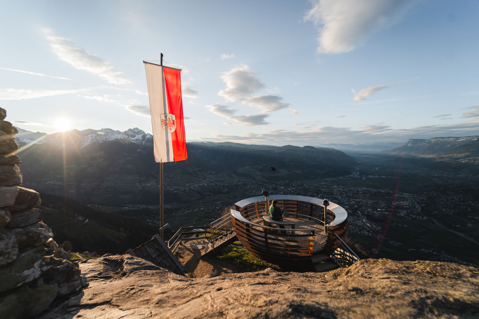 A panoramic view of the Merano region from the viewing platform at the top station of the Aschbach cable car. A South Tyrolean flag flutters in the evening light, whilst mountains, valleys and villages stretch as far as the eye can see – an impressive des
