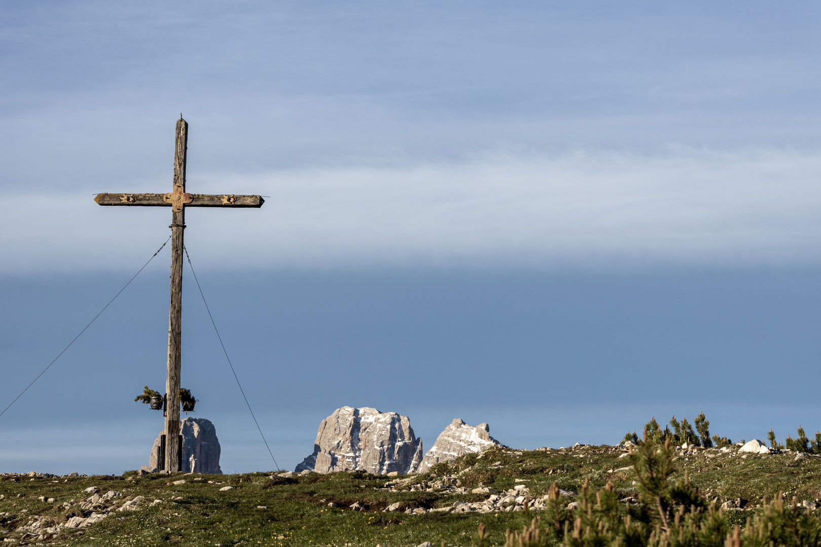 Ein schlichtes Gipfelkreuz steht auf einer felsigen Hochebene mit alpiner Vegetation. Im Hintergrund ragen markante, schneebedeckte Dolomitengipfel unter einem dramatischen Himmel mit dunklen Wolken auf, während warmes Sonnenlicht die Landschaft kontrastr