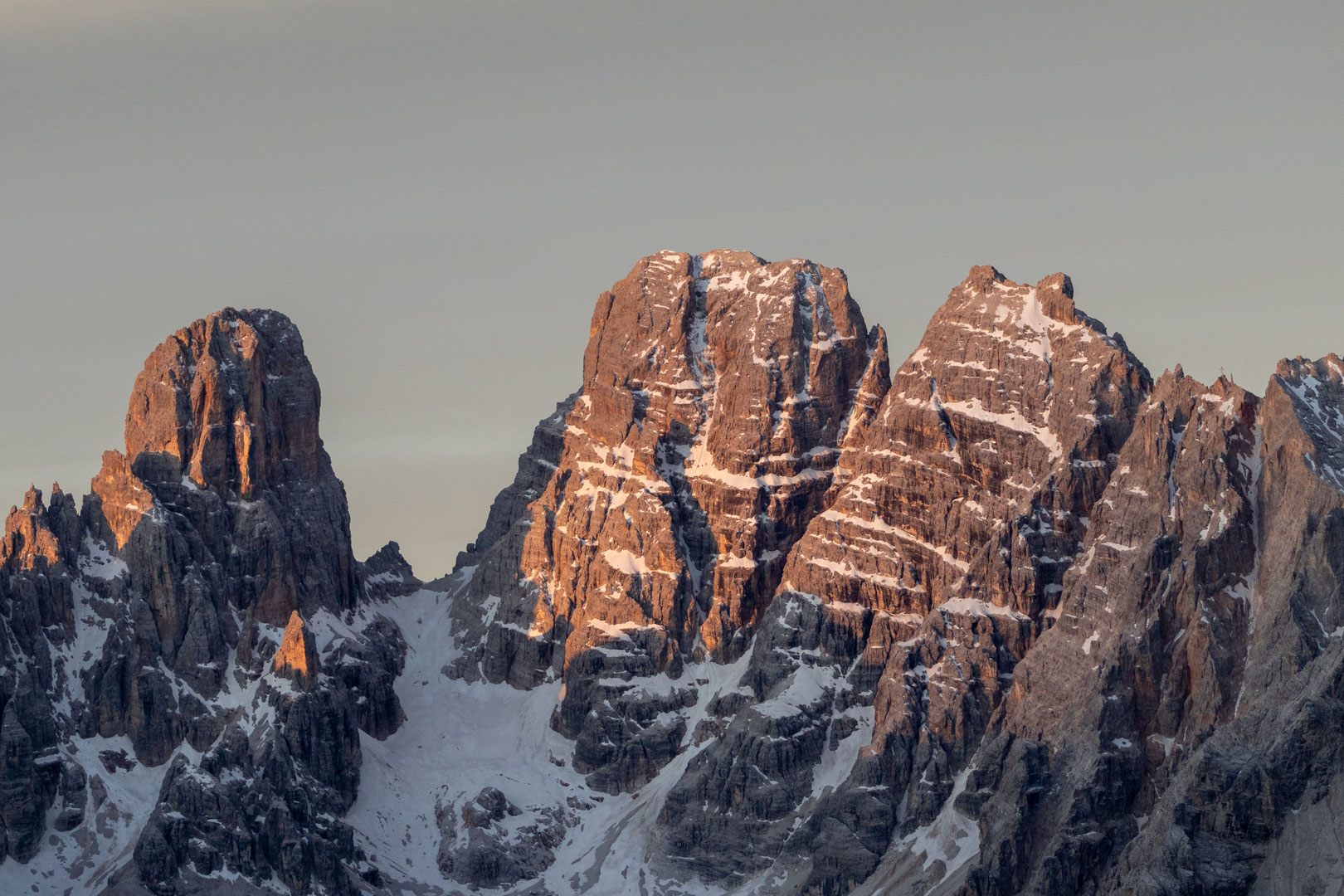 Nahaufnahme einer markanten Dolomitengruppe im warmen Licht der aufgehenden oder untergehenden Sonne. Die schroffen Felswände sind von Schneefeldern durchzogen und heben sich vor einem ruhigen, wolkenlosen Himmel in sanften Grautönen ab.
