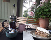 Kaffeegesdeck bestehend aus Kanne Kaffee, Becher und einem Stück Kucken auf einem Tablett auf einem Bistrotisch mit einem Geranientopf