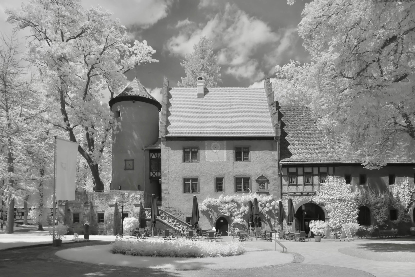 Das Gesindehaus von Schloss Aschach zeigt sich in Schwarz-Weiß mit Infraroteffekt. Das historische Gebäude ist umgeben von hell leuchtendem Baumbestand. Im Vordergrund sind Außenmöbel eines Cafés oder Museums zu sehen.