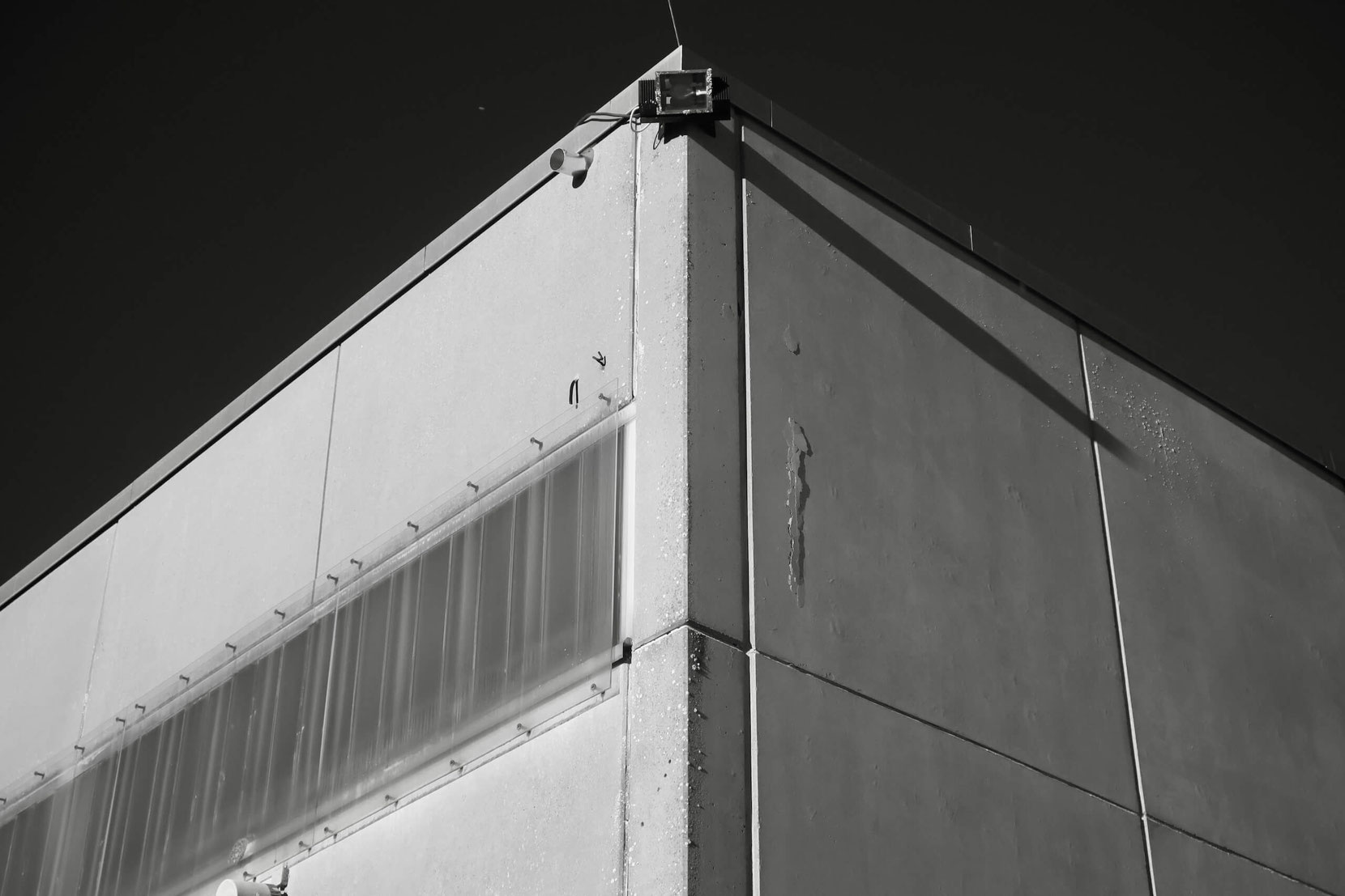 Schwarz-weiß-Aufnahme der Gebäudeecke eines ehemaligen Kasernengebäudes auf dem Gelände der Marshall Heights in Kitzingen. Zu sehen sind eine Betonfassade, Leitungen und ein montierter Außenstrahler.