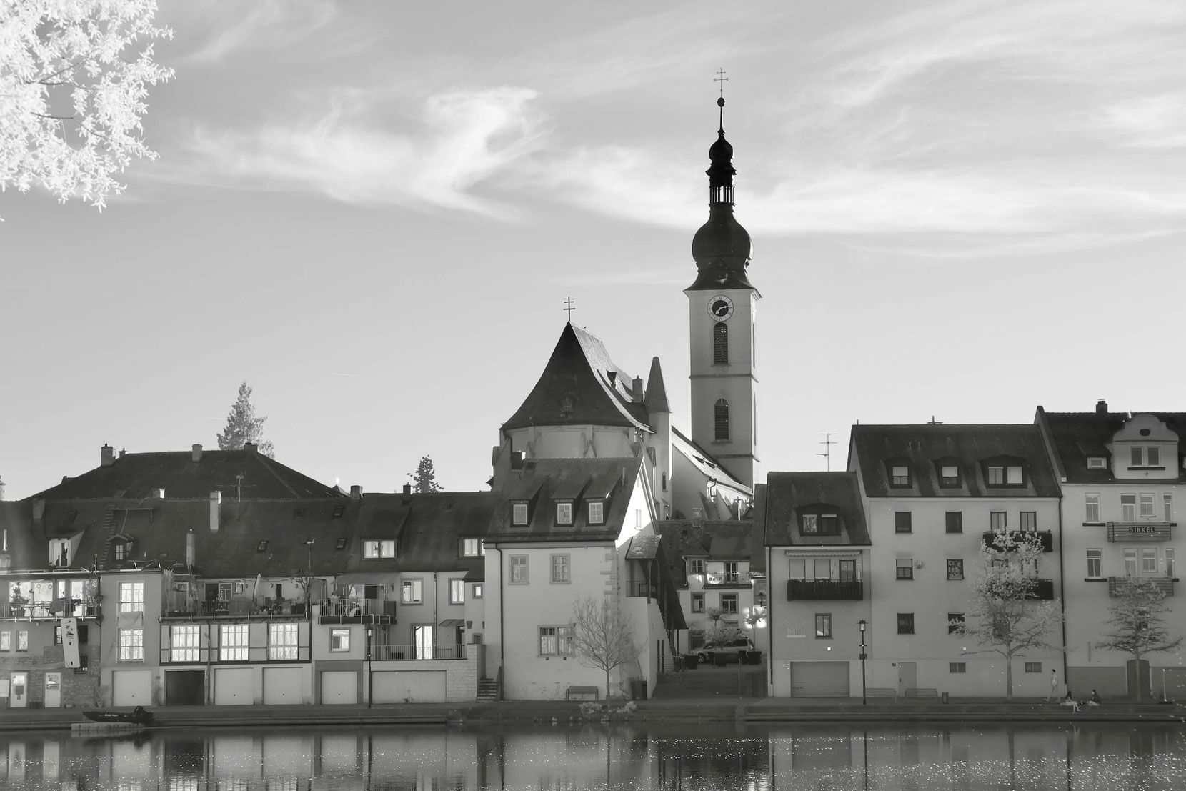 Die markante Silhouette der St.-Johannes-Kirche in Kitzingen. Ihr hoher Kirchturm mit Zwiebelhaube und Uhr ragt über die angrenzenden Häuser entlang des Mainufers hinaus, deren Fassaden sich im ruhigen Wasser spiegeln.