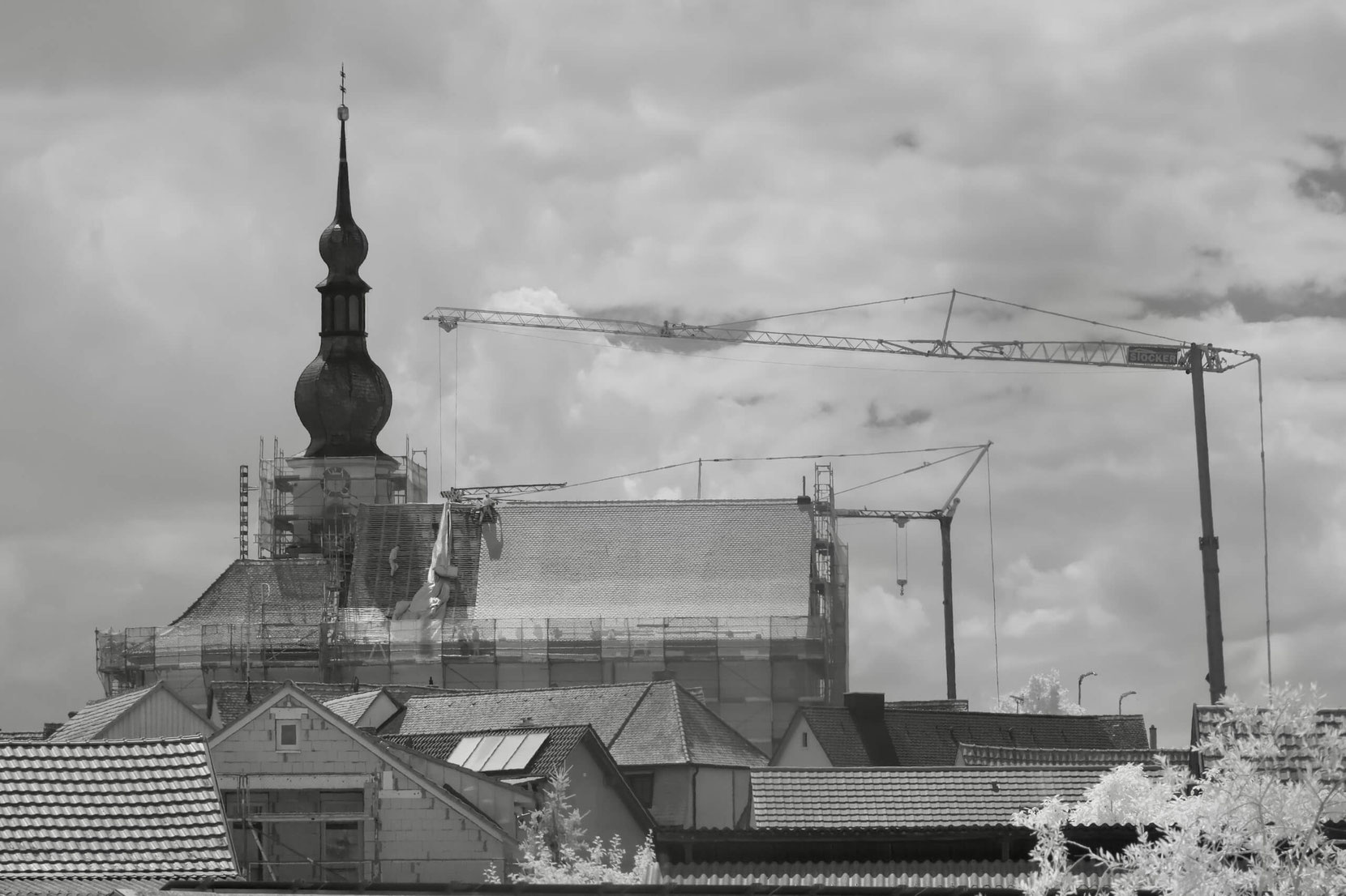 Das Bild zeigt eine Kirche mit eingerüstetem Turm während Sanierungsarbeiten, im Vordergrund Wohnhäuser und im Hintergrund mehrere Baukräne.