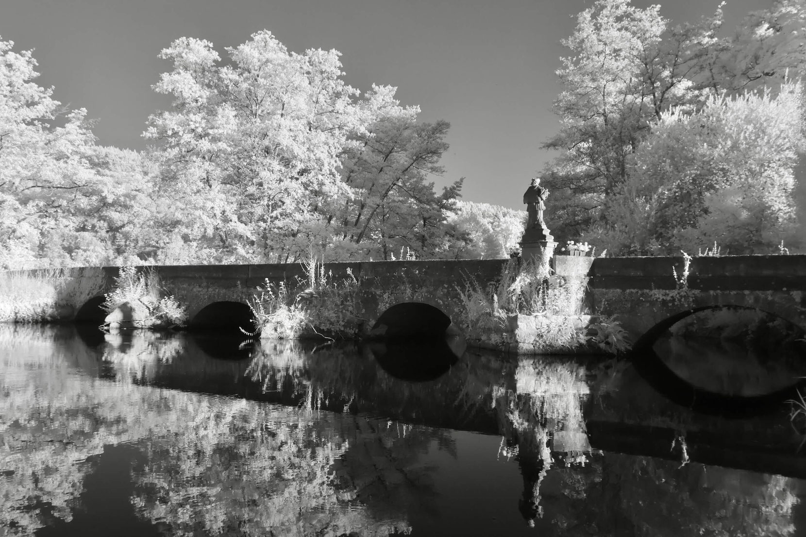 Eine historische Steinbrücke mit Bögen überquert die Sinn bei Schaippach. Eine Skulptur steht am Geländer, während die Bäume durch die Infrarotaufnahme in hellem Weiß erscheinen. Die Szene ist in Schwarz-Weiß gehalten, mit Spiegelungen im Wasser.