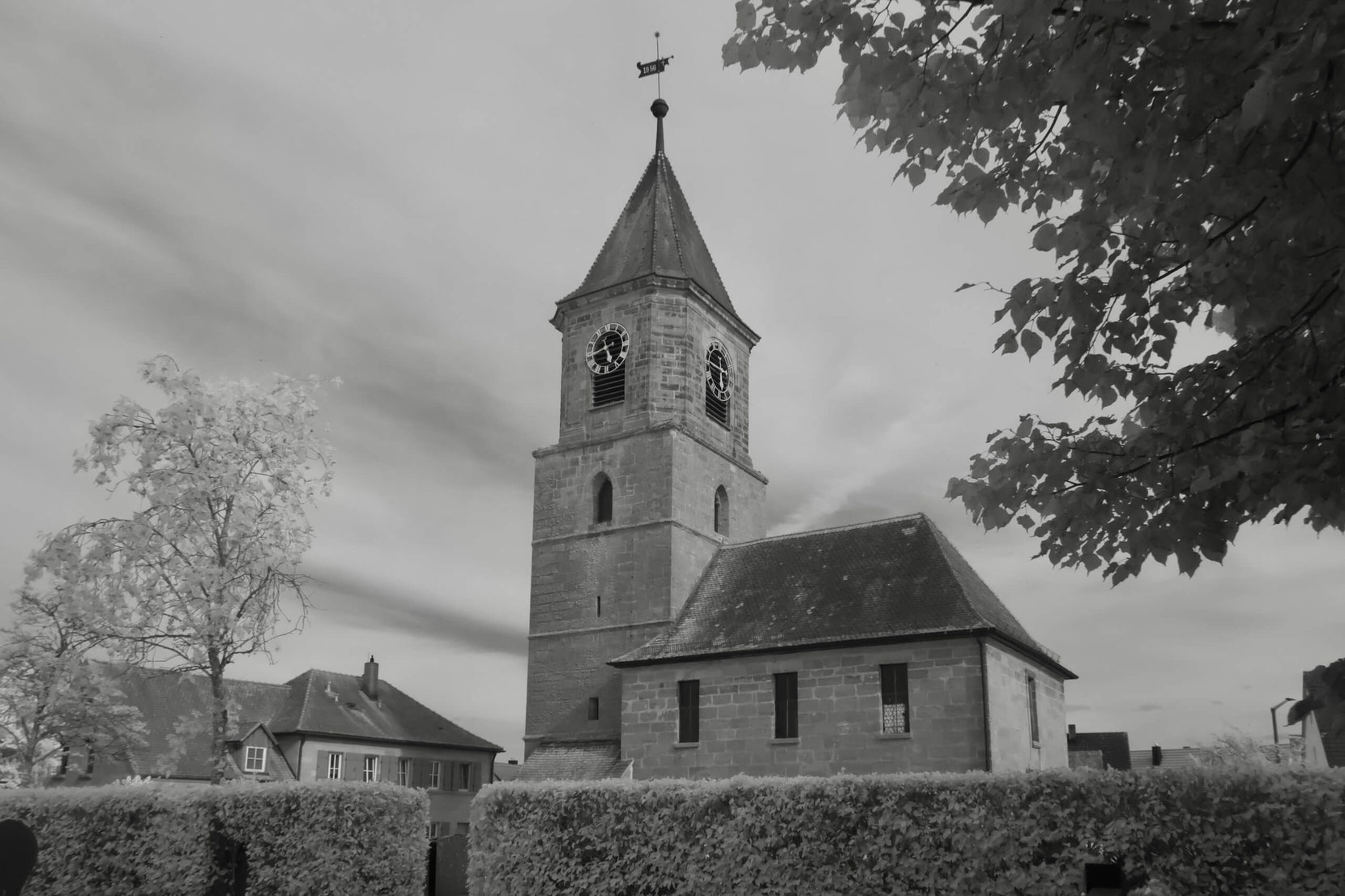 Die Evang.-Luth. Kirche St. Marien in Wallesau, Roth, mit ihrem markanten Turm ist in dieser Schwarz-Weiß-Infrarotaufnahme zu sehen. Die hell leuchtenden Bäume und die Hecke im Vordergrund verleihen der Szene eine besondere, leicht surreale Ästhetik.