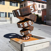Public sculpture by Paolo Grassi titled Thiago, corten steel numeric structure in front of a modern architectural building.