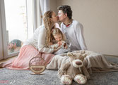 das Foto zeigt eine Familie in ihrem Wohnzimmer. sie sitzen auf dem Boden. Die Eltern küssen sich und die Tochter im Kitaalter küsst das Neugeborene Baby. Die Farben sind im Bohostil gehalten. 