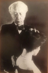 medium closeup of man (F.M. Alexander) standing with hand on head and upper back of seated female student 